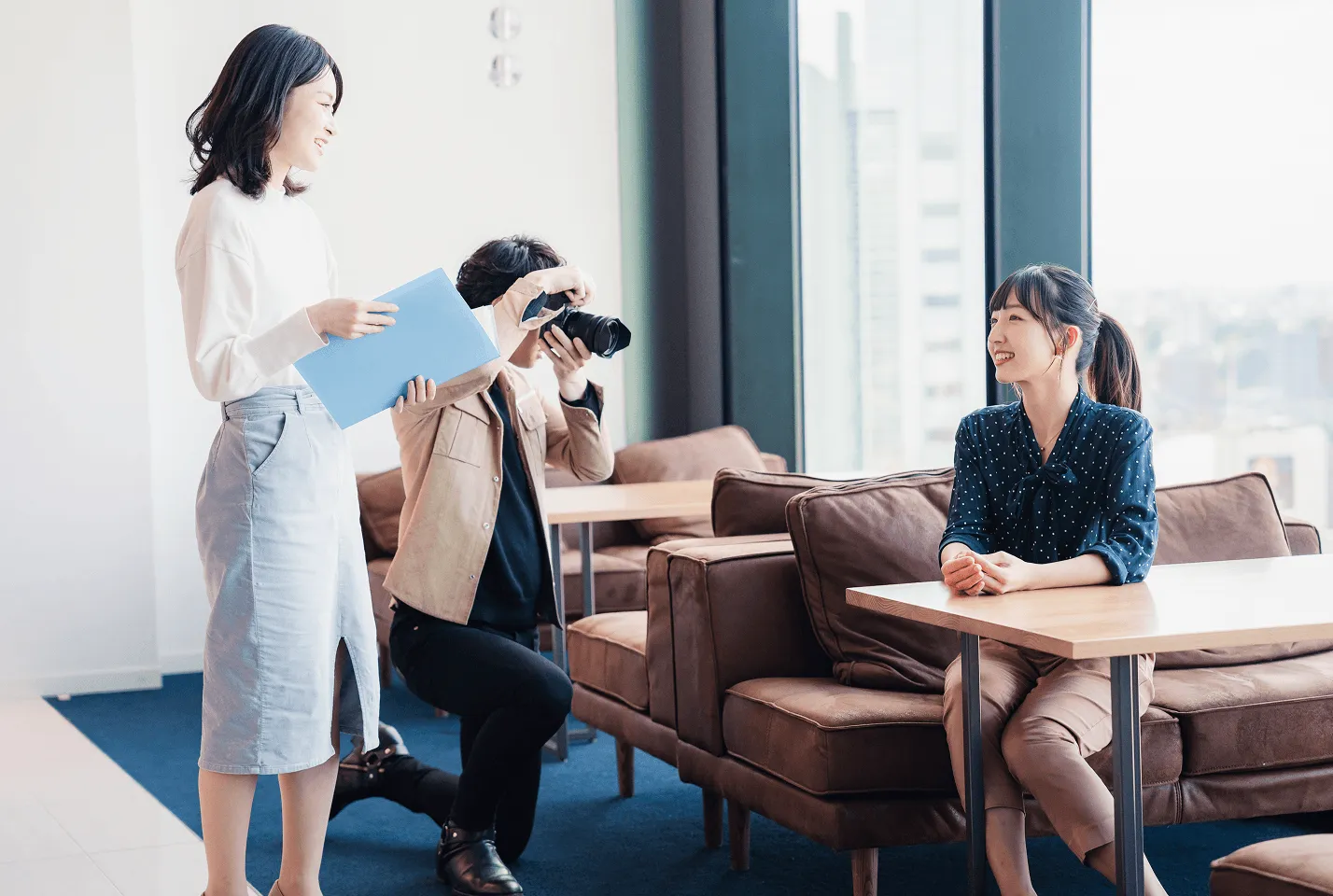 窓際のラウンジで、資料を持つ人とカメラを構える人、ソファに座る人が向き合う様子